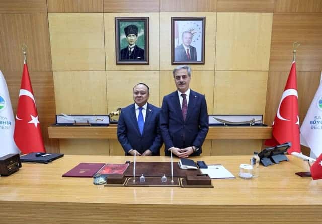 Görselde iki yönetici, belediye odasında duruyor, arka planda Türk bayrakları ve resmi portreler bulunuyor.